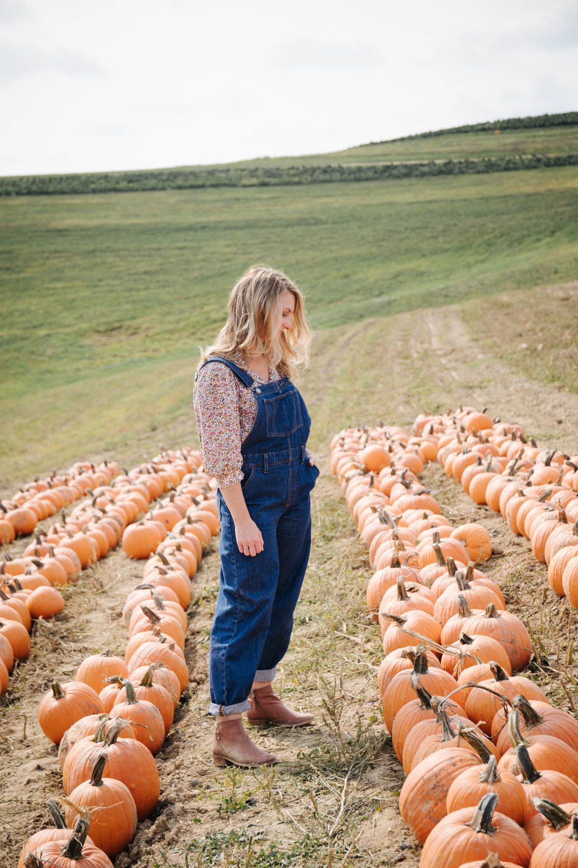 Allyn Lewis shares cute outfits to wear to a pumpkin patch wearing a casual fall outfit with denim overalls and brown ankle booties. 