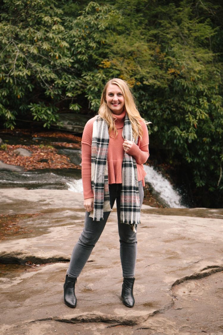 women hiking by a waterfall wearing a grey jeans outfit styled with black ankle boots, pink sweater, and plaid scarf.