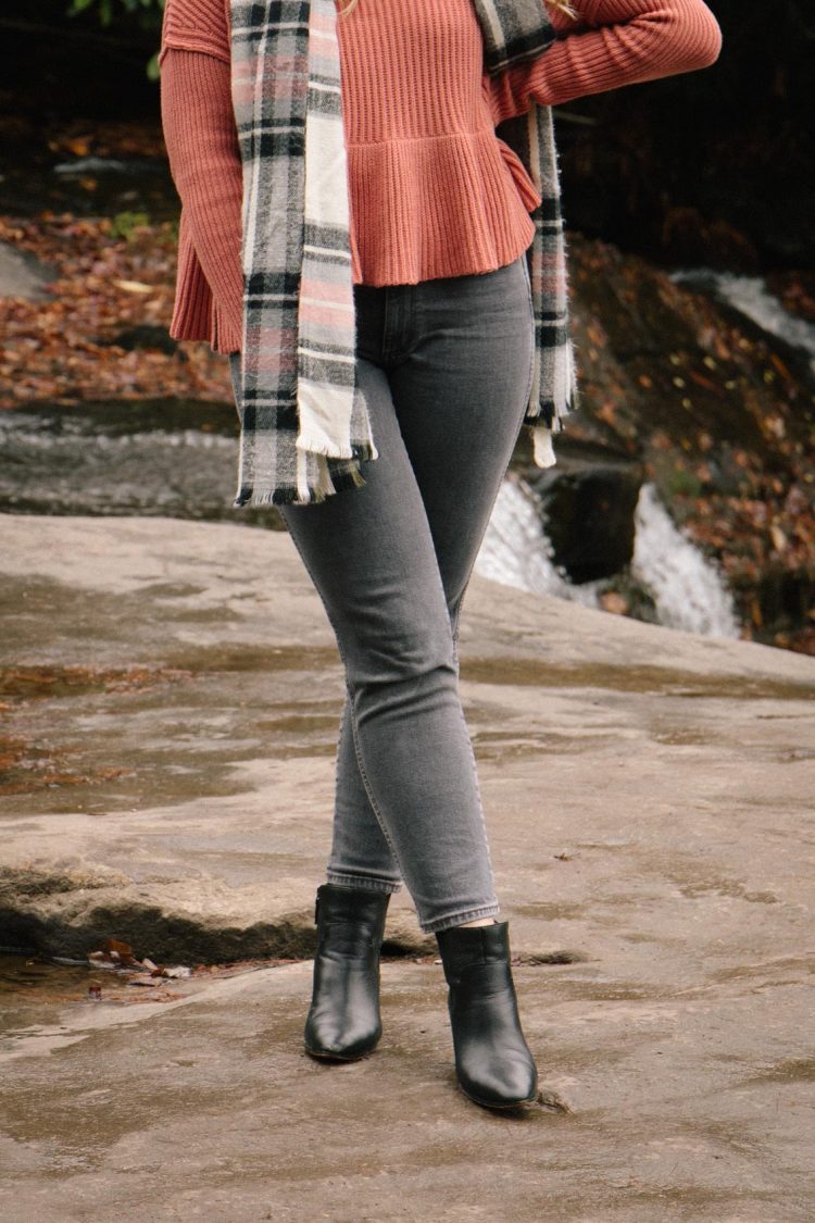 Women's casual winter outfit styled with grey high rise jeans, a pink peplum sweater, plaid scarf, and grey ankle boots.