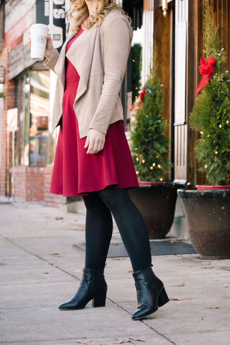Classy winter outfit featuring a women wearing a red fit and flare dress, tights, faux suede jacket, and black ankle boots.