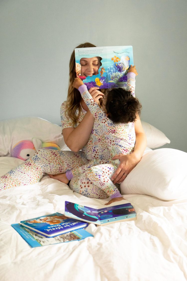 Woman and toddler playing on a white bed reading books wearing matching bamboo pajamas in a floral print from Little Sleepies pajamas.
