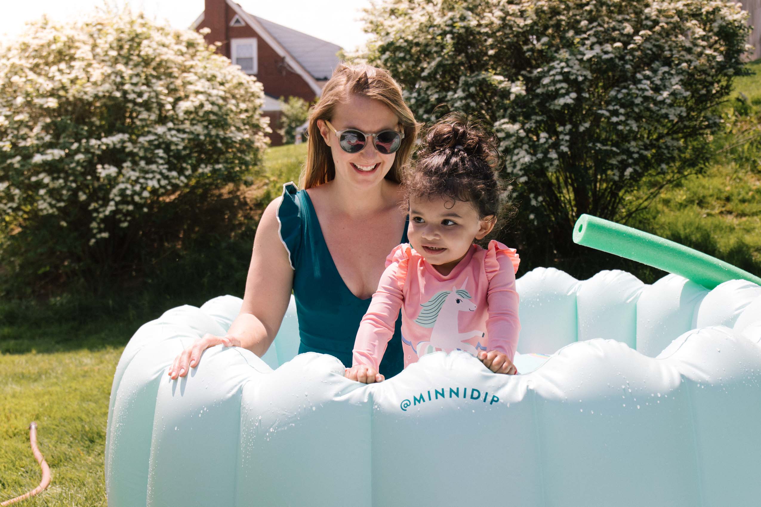 Woman in a teal one piece Summersalt bathing suit with a toddler in an inflatable kiddie pool. 