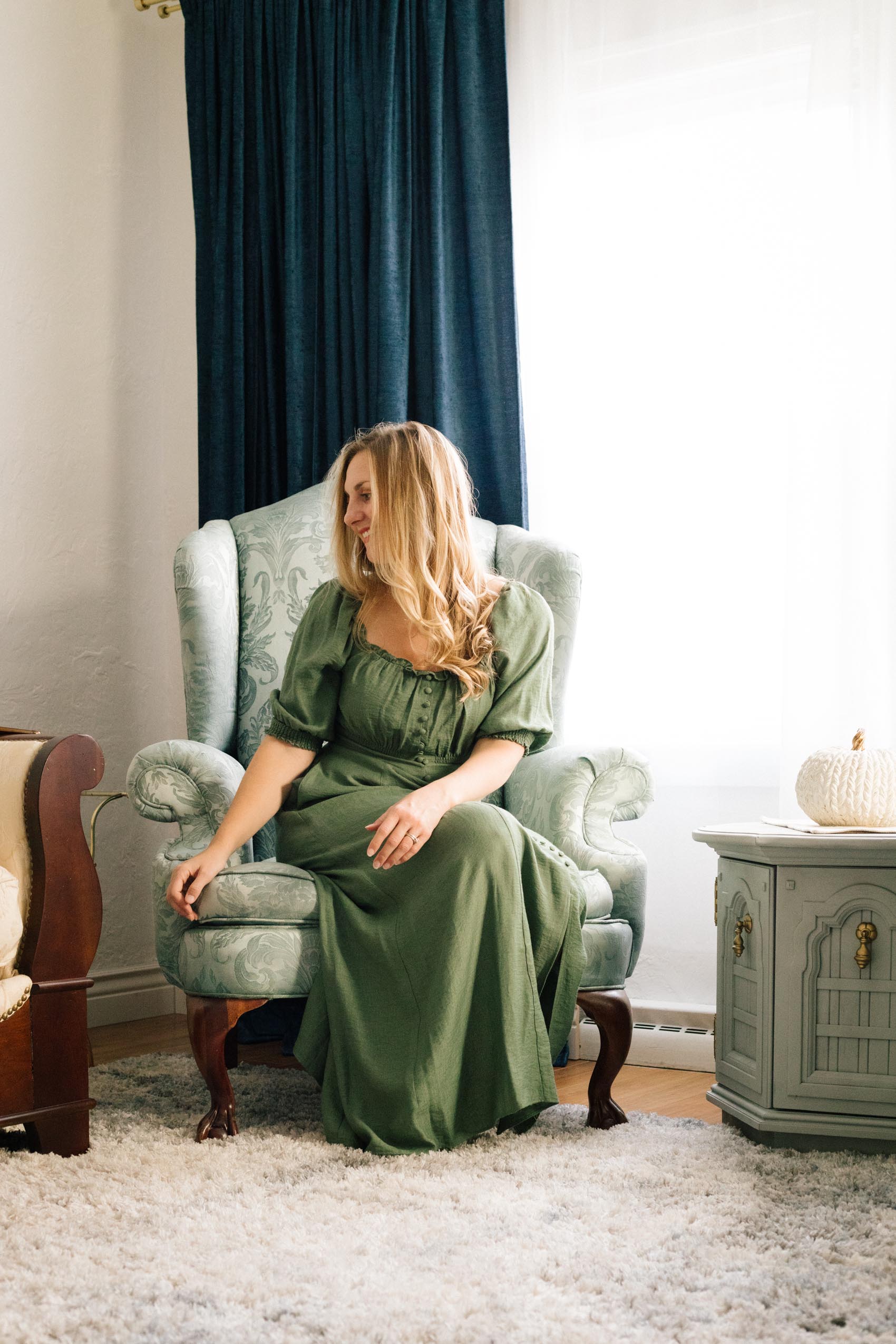Women in a green dress sitting on a vintage blue wing back chair in front of dark blue curtains