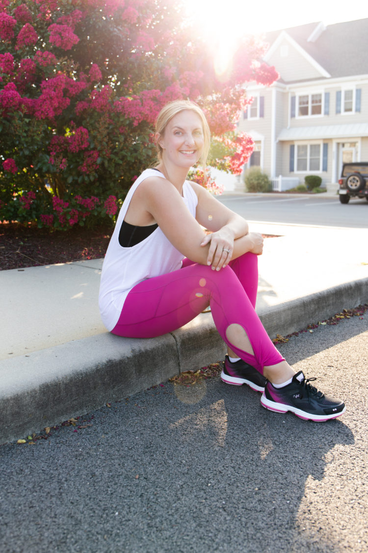 Woman setting on a curb wearing back Ryka sneakers, a white tank top, and pink activewear leggings.
